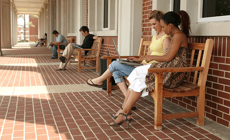 College students sit outside on benches along a wall while studying.