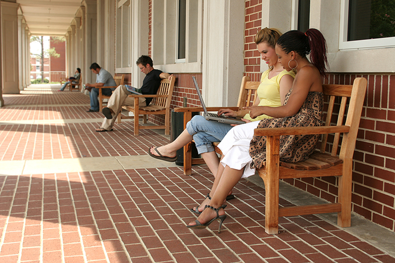College students sit outside on benches along a wall while studying.