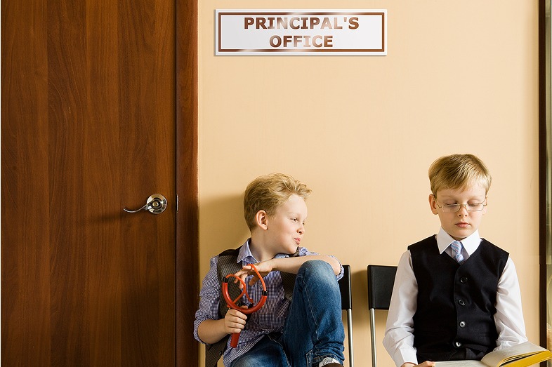 Two schoolboys sitting next to principal's office. They have different behavior and clothes.