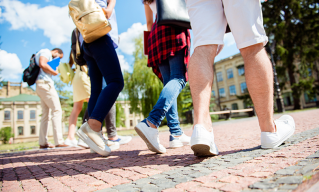 Low angle rear view shot of six students' feet. They are walking outside near a college building on a sunny summer day.