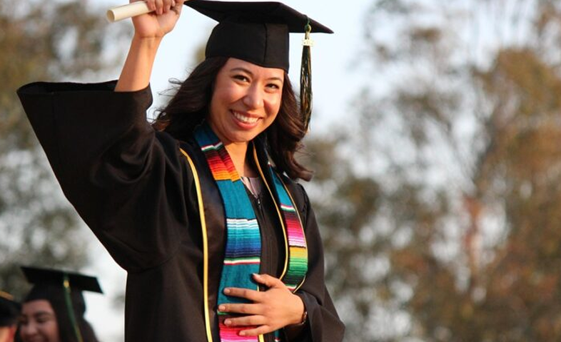 A Latina college graduate stands in her cap and gown, raising her diploma in the air.