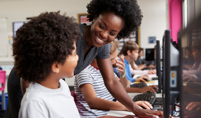A female teacher helps a boy student who is working on a computer.
