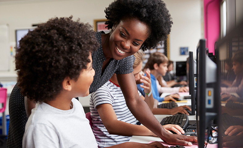 A female teacher helps a boy student who is working on a computer.