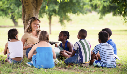 A young woman teacher reads a book to boys and girls at a park.