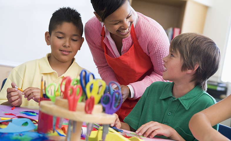 A female teacher speaks to two young male students as they work on art projects.