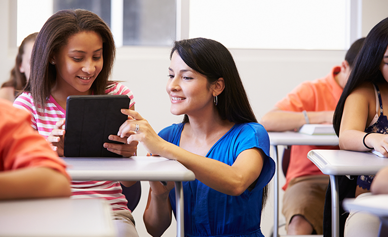 A female teacher stoops down near a student's desk to help her with an assignment.